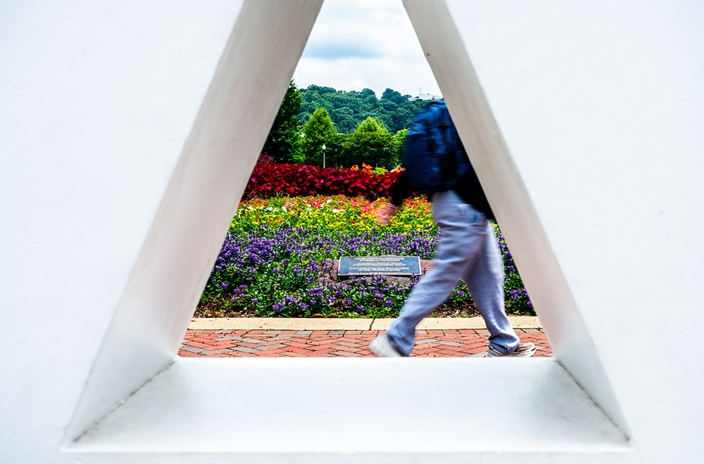 The back and legs of a student wearing jeans and a blue backpack are seen through the A in the large U A B letters on the Campus Green, with flowers and trees visible behind the student