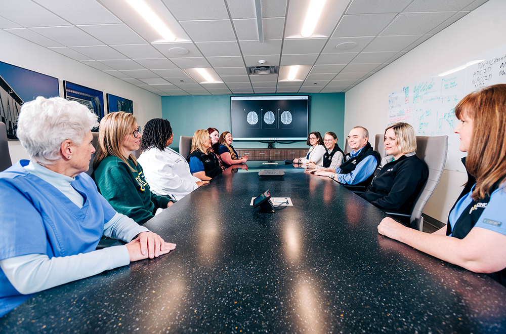 Members of the care team at the UAB Brain Aging and Memory Clinic gather around a conference table, with a screen showing mock brain scans behind them