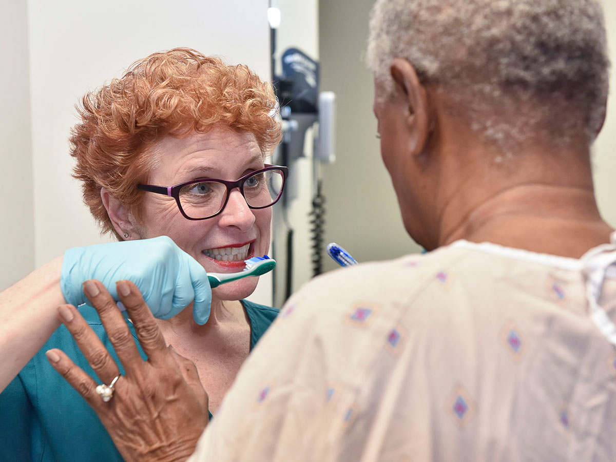 Rita Jablonski holds a toothbrush in her gloved hand and demonstrates brushing to a patient, wearing hospital gown, with back to camera