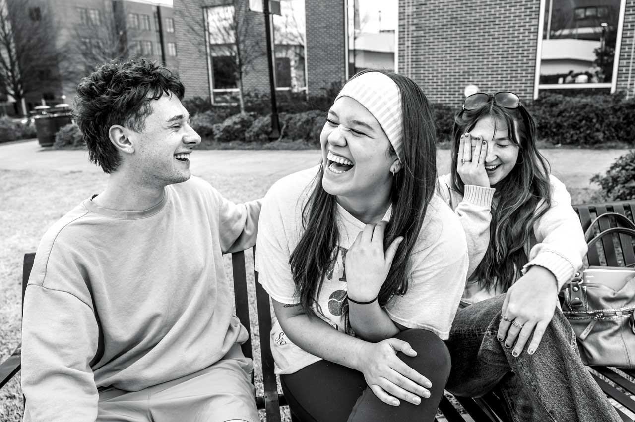 Three students sit together outside, laughing and talking near a brick building.