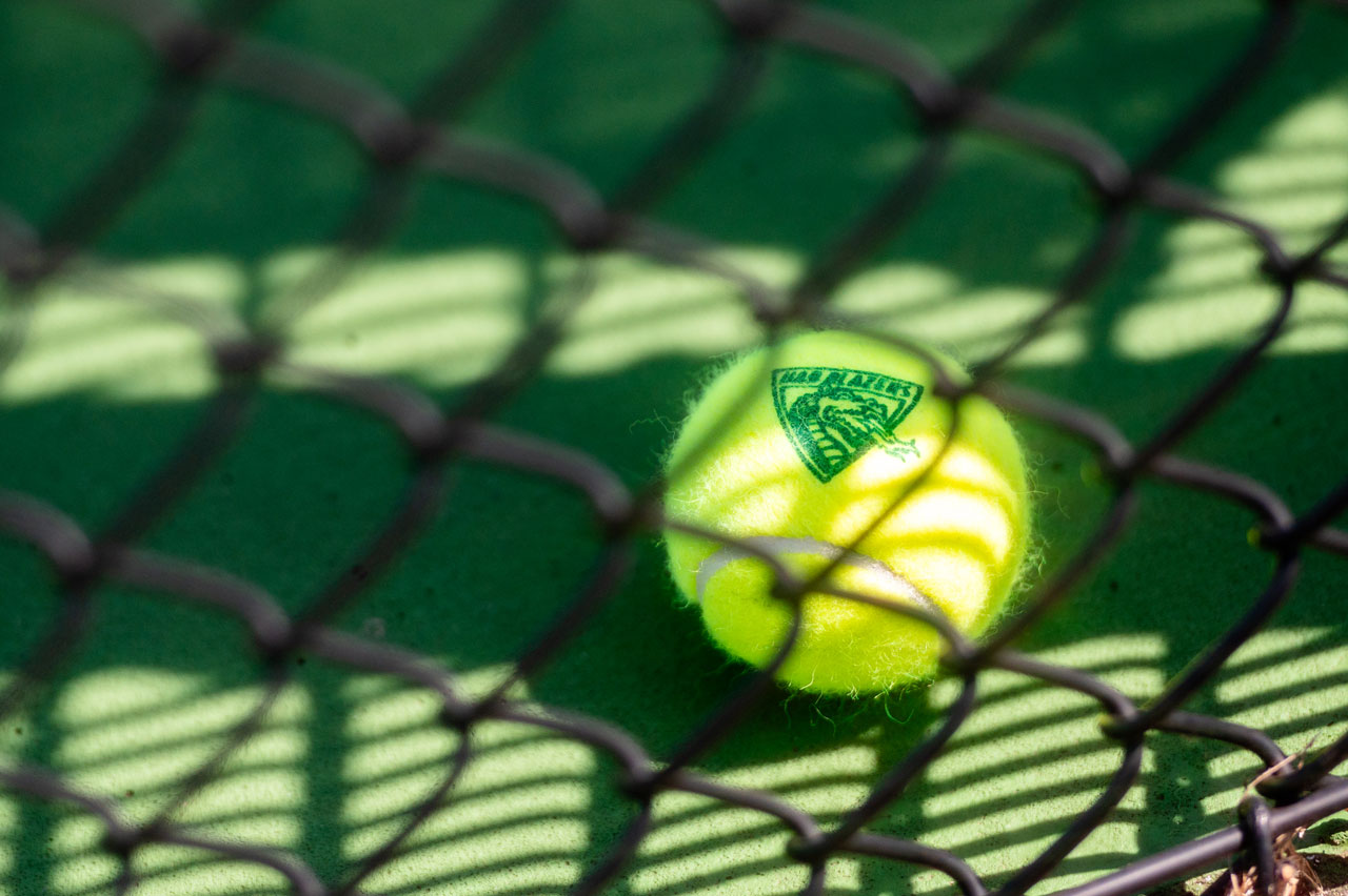 A tennis ball stamped with the UAB Blazers logo is embedded in a segment of a chain link fence.