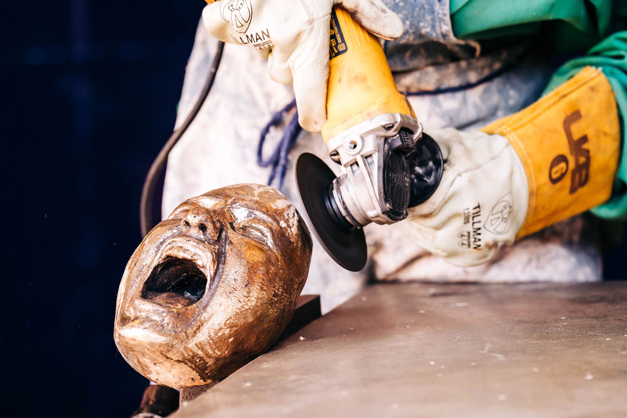 Gloved hands use a sander to shape a wooden mask.